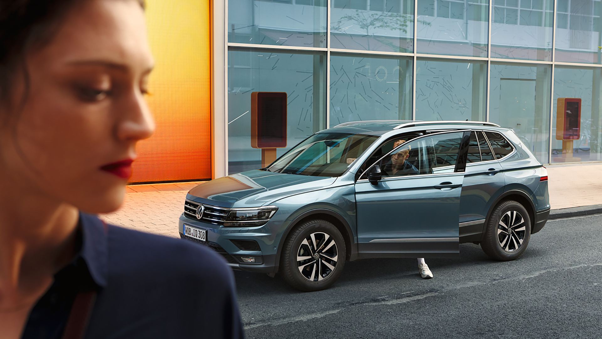 Uomo esce dalla sua Volkswagen Touran, in primo piano una donna vista di lato